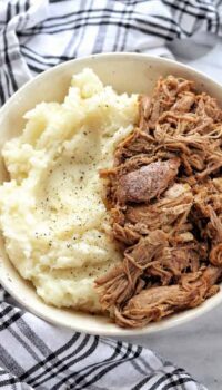 A bowl of pulled pork next to mashed potatoes with black pepper on top.