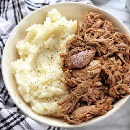A bowl of pulled pork next to mashed potatoes with black pepper on top.