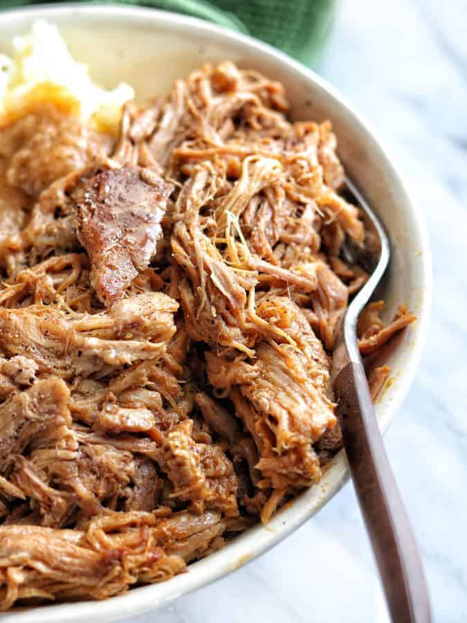 instant pot pork shoulder recipe shown close up in a white bowl with a fork on a white marble surface. 