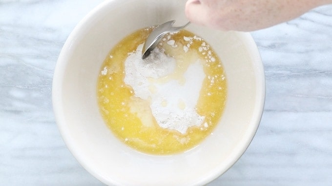 Mini tarts recipe being made showing the crust ingredient of melted butter, flour, and sugar in a white bowl being stirred on a white marble surface.