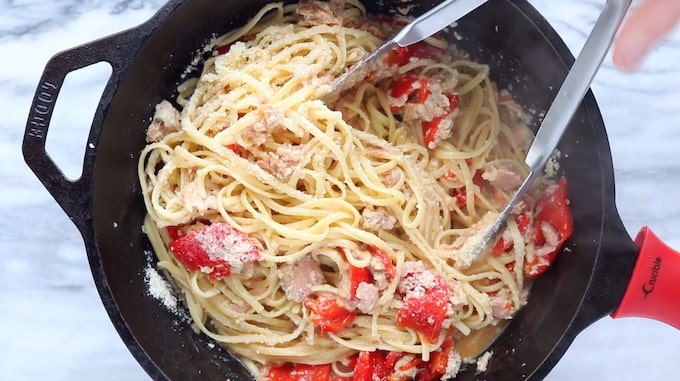 A tuna pasta recipe shown in a black cast iron pan with pasta and parmesan cheese being blended to gather with the tinned tuna mixture.