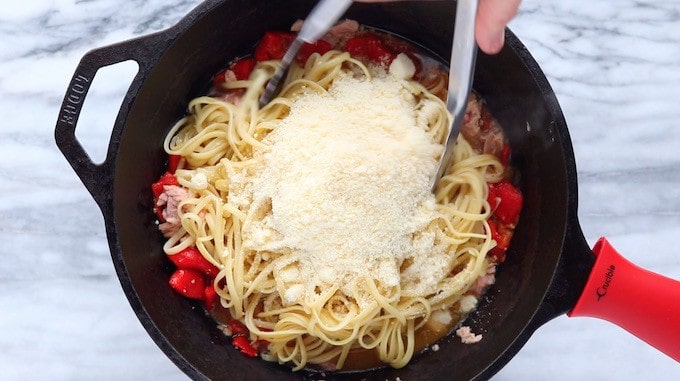 A tuna pasta recipe shown in a black cast iron pan with pasta and parmesan cheese on top ready to be blended to gather with the tinned tuna mixture.