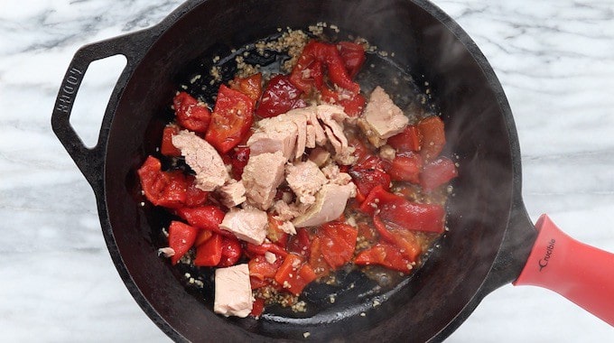 A tuna pasta recipe being made showing albacore tuna added to the recipe in a black cast iron pan with roasted red peppers. 