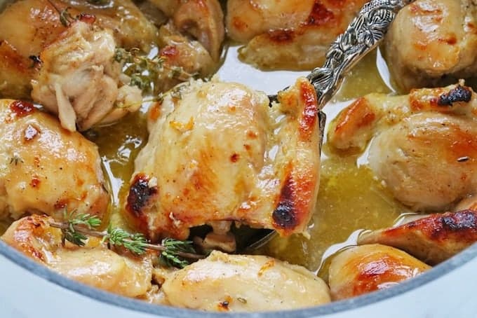 Boneless Skinless Chicken thighs stovetop shown in a white pot. Chicken thighs are browned and shown in juice with fresh herbs close up.
