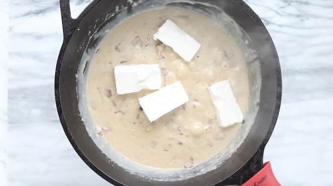 Creamy Chicken and Bacon Pasta recipe being made, showing creamy sauce in a black cast iron pan with cream cheese chunks placed into the sauce.