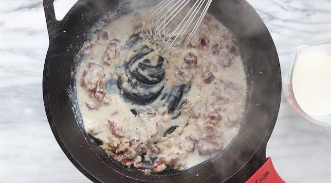 Creamy Chicken and Bacon Pasta recipe being made, showing bacon, garlic, and milk being whisked into flour in a black cast iron pan.