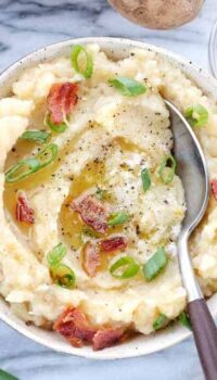 A bowl of colcannon is shown in a white bowl topped with bacon, butter, and green onions.