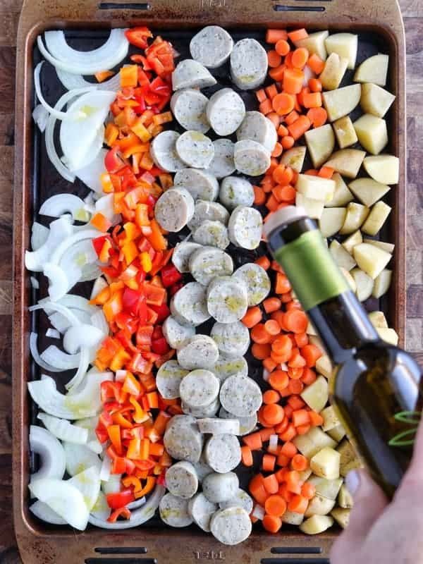 al fresco chicken sausage shown on a baking sheet with potatoes, carrots, peppers, and onions with olive oil being drizzled onto it. 