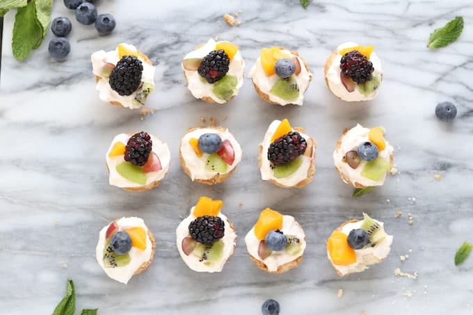 Summer fruit tart shown in 3 rows of 4 topped with fruit with mint and blueberries around the outside on white marble.