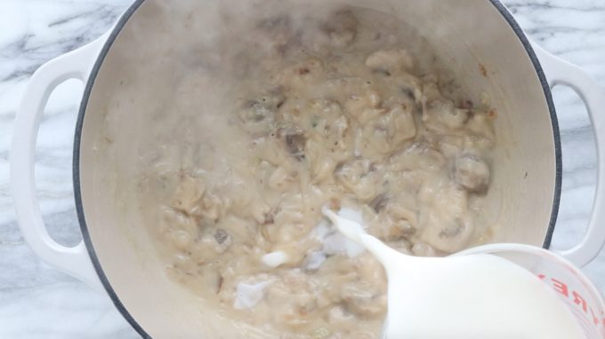 milk is being whisked into the onion, mushroom, butter and flour mixture in a white stockpot on a marble table.