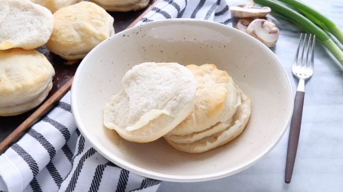 biscuits are shown in a white bowl with a fork next to it and a baking tray of freshly bakes biscuits in the back.