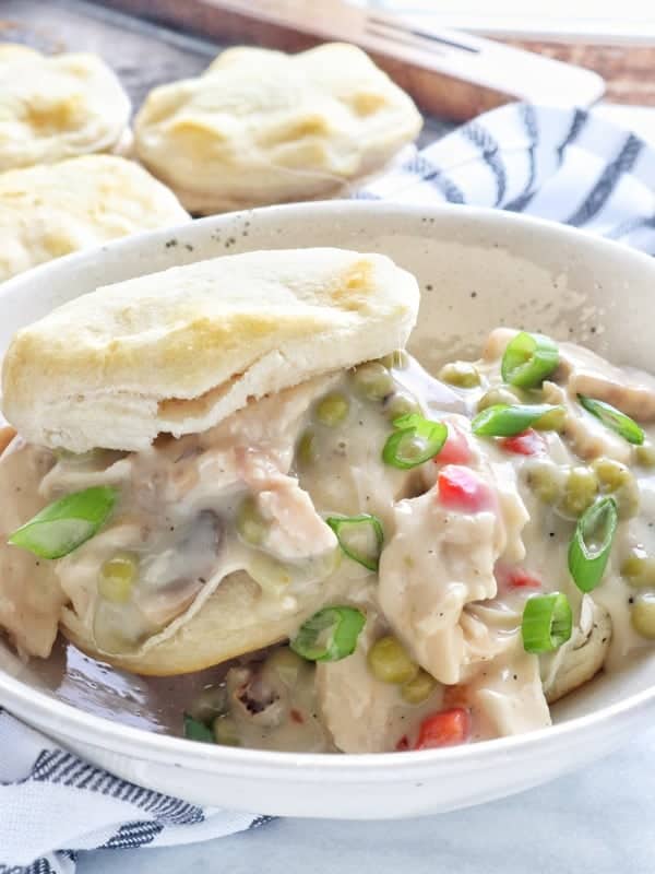 Creamy chicken a la king in a white bowl spilling from a biscuit with peas, mushrooms with more biscuits in the background.