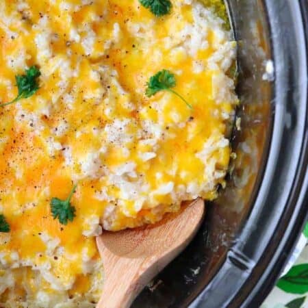 Cheesy Potatoes shown in a black crockpot with a wooden spoon.