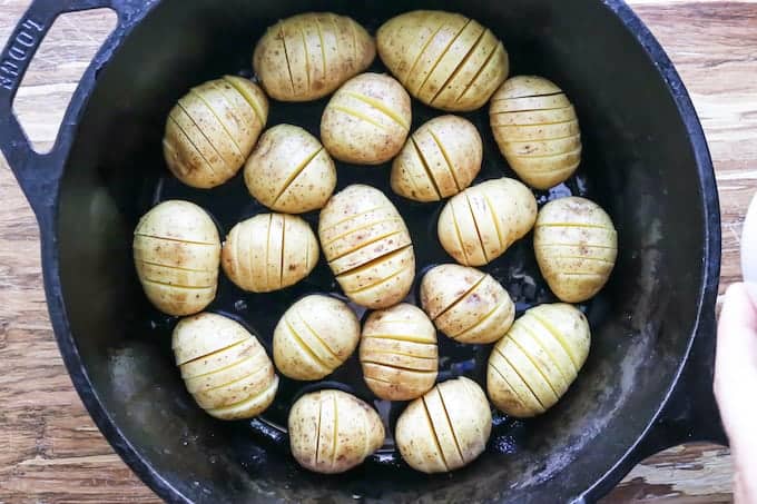 Hasselback potatoes with cheese being made in a black cast iron skillet showing the potatoes with slices on a wooden surface.
