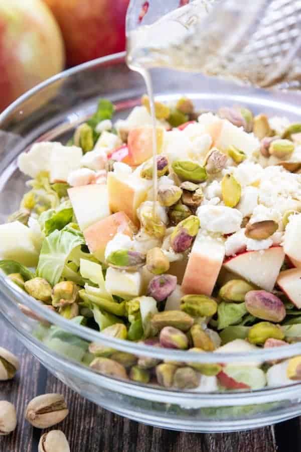 A Brussels sprouts salad in a glass mixing bowl topped with pistachios, apple chunks and feta cheese. 