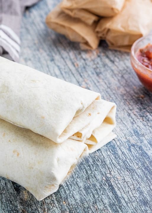 Refried Bean Burrito, a vegetarian camping recipe showing wrapped and unwrapped burritos, a towel and glass bowl of salsa all on a wooden surface.