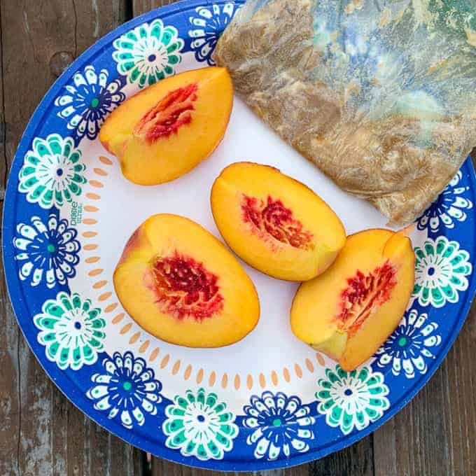 easy campfire desserts showing a sliced peach with butter and sugar mixed in a ziplock bag on a disposable plate on a picnic table