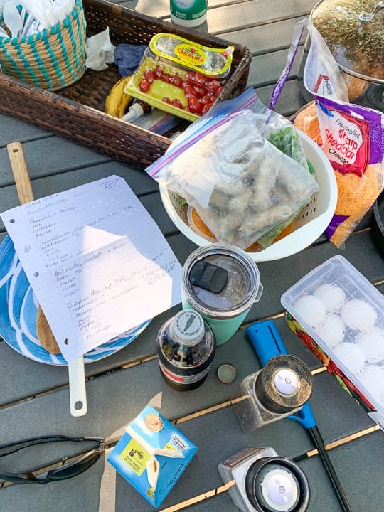 Camping menu shown on a picnic table next to breakfast items like sausage, eggs, and cheese. 