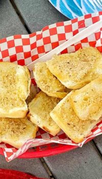Garlic toast is in a red plastic serving bowl with red and white checkered paper on a picnic table.