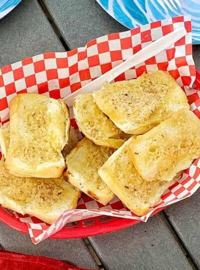 Garlic toast is in a red plastic serving bowl with red and white checkered paper on a picnic table.