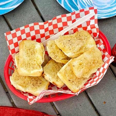 Garlic toast is in a red plastic serving bowl with red and white checkered paper on a picnic table.