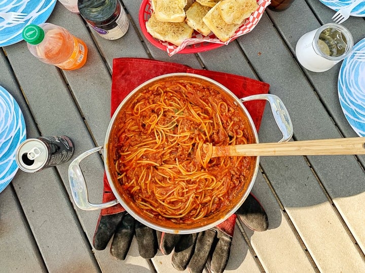 One pot camping meals: spaghetti shown in a stainless steel pot with a wooden spoon next to garlic bread on a picnic table.