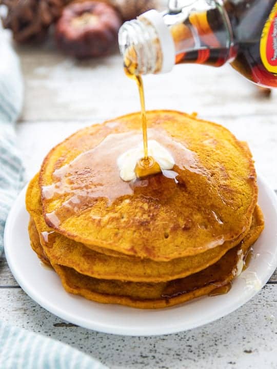 Healthy Pumpkin Pancakes, a pumpkin spice pancake recipe showing maple syrup being pour over the top of stacked pancakes on a white plate.