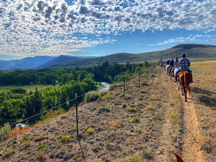 riders on horses on a path next to a hill where a river is running down below with mountains in the distance and bright sun with clouds in the sky.