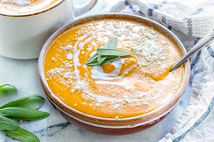 butternut squash soup shown in a bowl drizzled with coconut milk and fresh sage leaves on a white surface.
