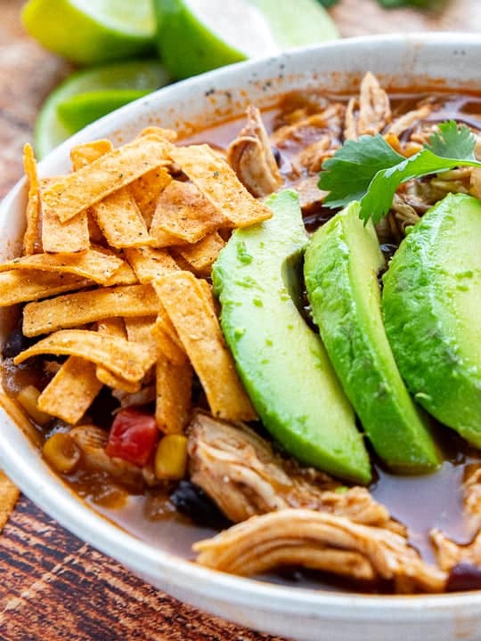 A chicken soup in a white bowl with sliced avocados and tortilla chips.
