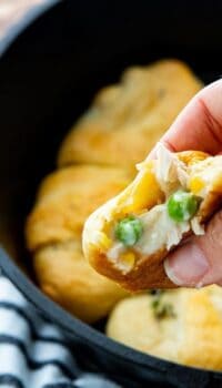 A hand is holding a chicken pot pie filled crescent roll with more crescents in a skillet blurred in the background.