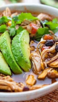 Chicken tortilla soup in a white bowl showing chicken chunks, corn, and beans with fresh avocados on top and a spoon.