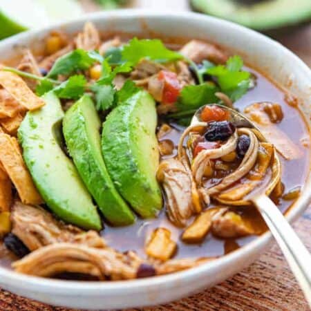 Chicken tortilla soup in a white bowl showing chicken chunks, corn, and beans with fresh avocados on top and a spoon.