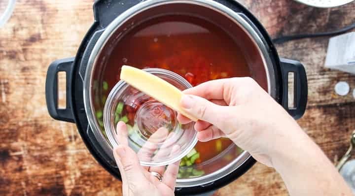 a top down view of an instant pot showing vegetables and beans with stock with a hand adding a parmesan rind to the pot.