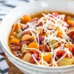 Minestrone soup in a white bowl packed with bean, carrots and celery with shredded cheese on top.