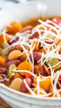 Minestrone soup in a white bowl packed with bean, carrots and celery with shredded cheese on top.