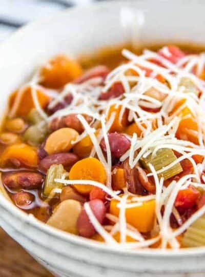 Minestrone soup in a white bowl packed with bean, carrots and celery with shredded cheese on top.