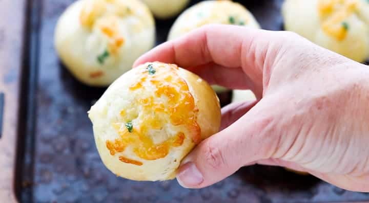 Parmesan Rhodes Rolls, a frozen dinner rolls recipe, showing Rhodes rolls on a baking sheet with a hand holding a dinner roll.