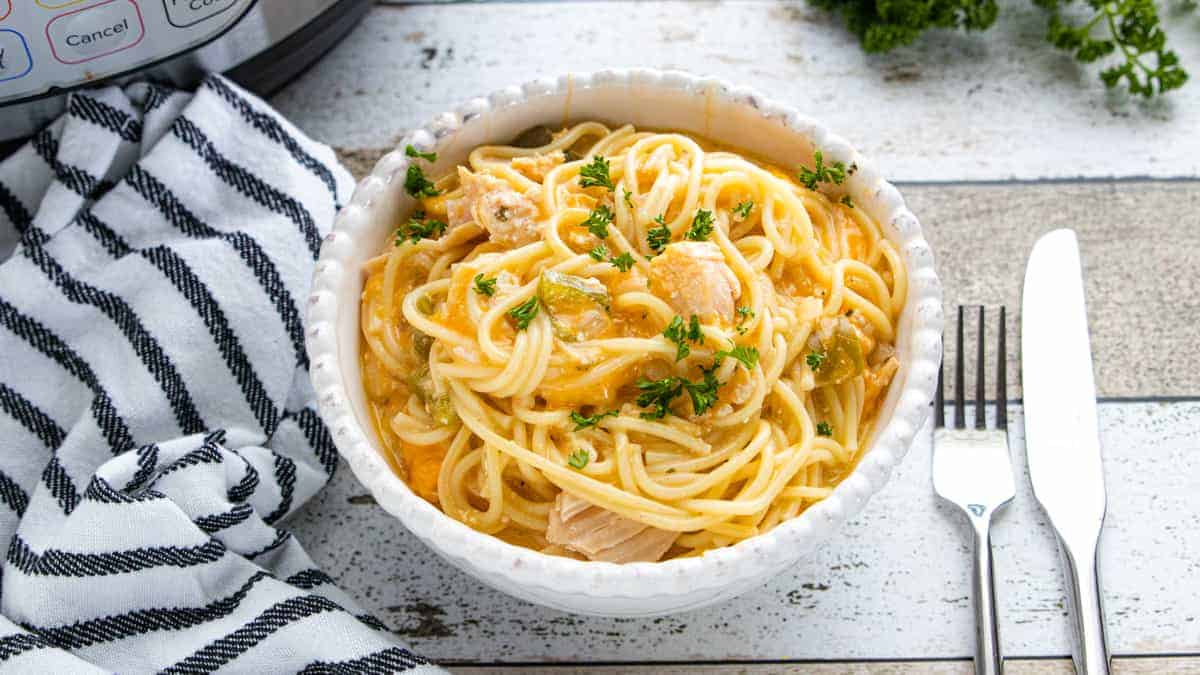 a white bowl filled with creamy chicken spaghetti and a fork and knife on the side.