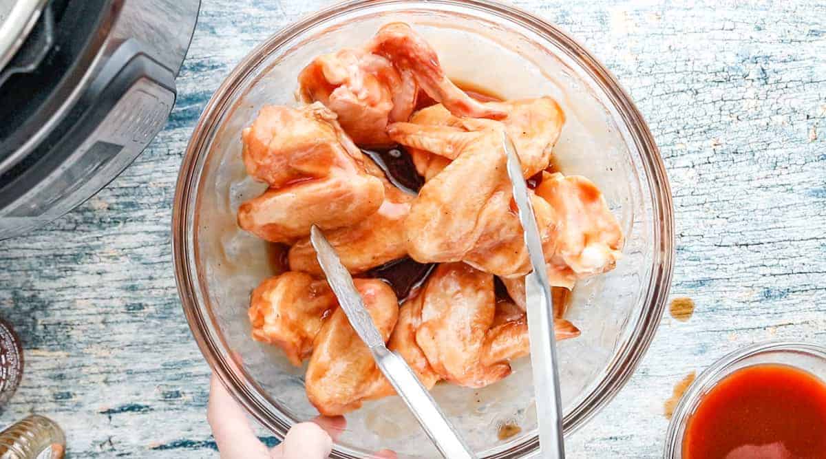  a large glass bowl filled with uncooked chicken with tongs coating the chicken in bbq sauce.