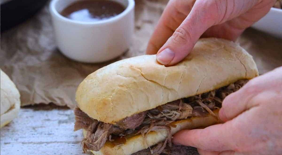 two hands picking up a French dip sandwich with a small bowl of au jus on a wooden countertop.
