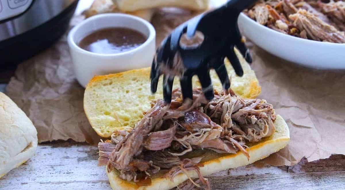 shredded rump roast being placed on a toasted French roll with a side of au jus next to the roll.