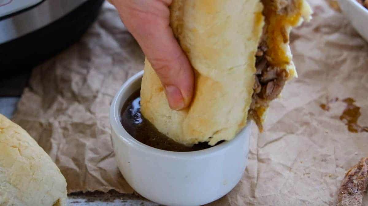a hand dipping a French dip sandwich into a small bowl of au jus.