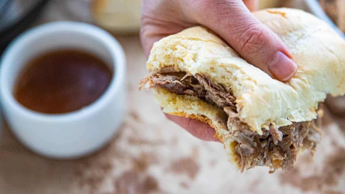 a hand holding a French dip sandwich with a bowl of au jus on a countertop.