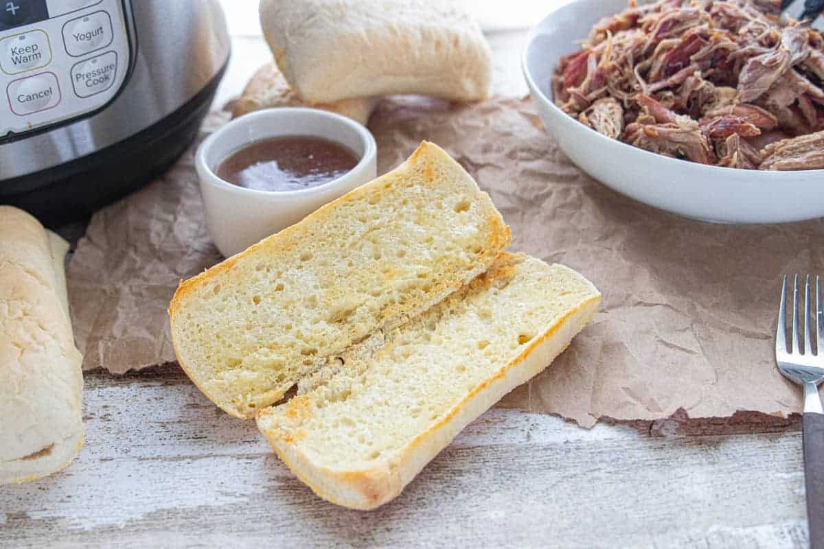  a toasted French roll, a small bowl of au jus, an instant pot and a large bowl of shredded beef on top of a wooden countertop.