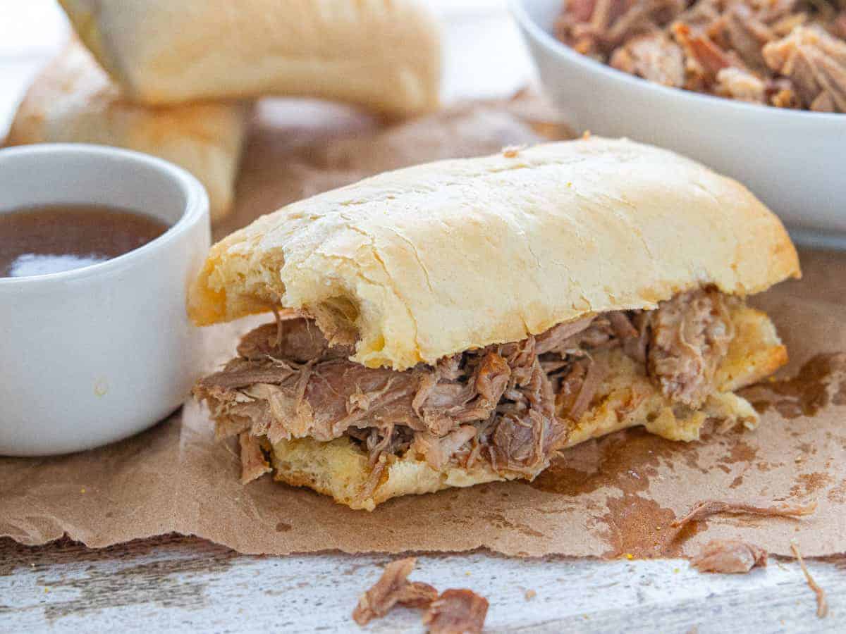 a French dip sandwich next to a small bowl of au jus with a large bowl of shredded beef all on top of a wooden countertop.