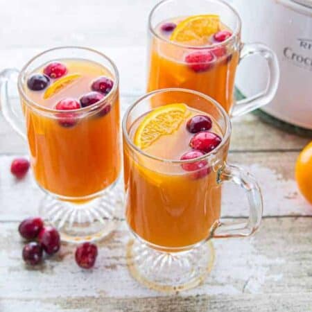 three glasses filled with apple cider next to a small crockpot, fresh cranberries, and oranges.