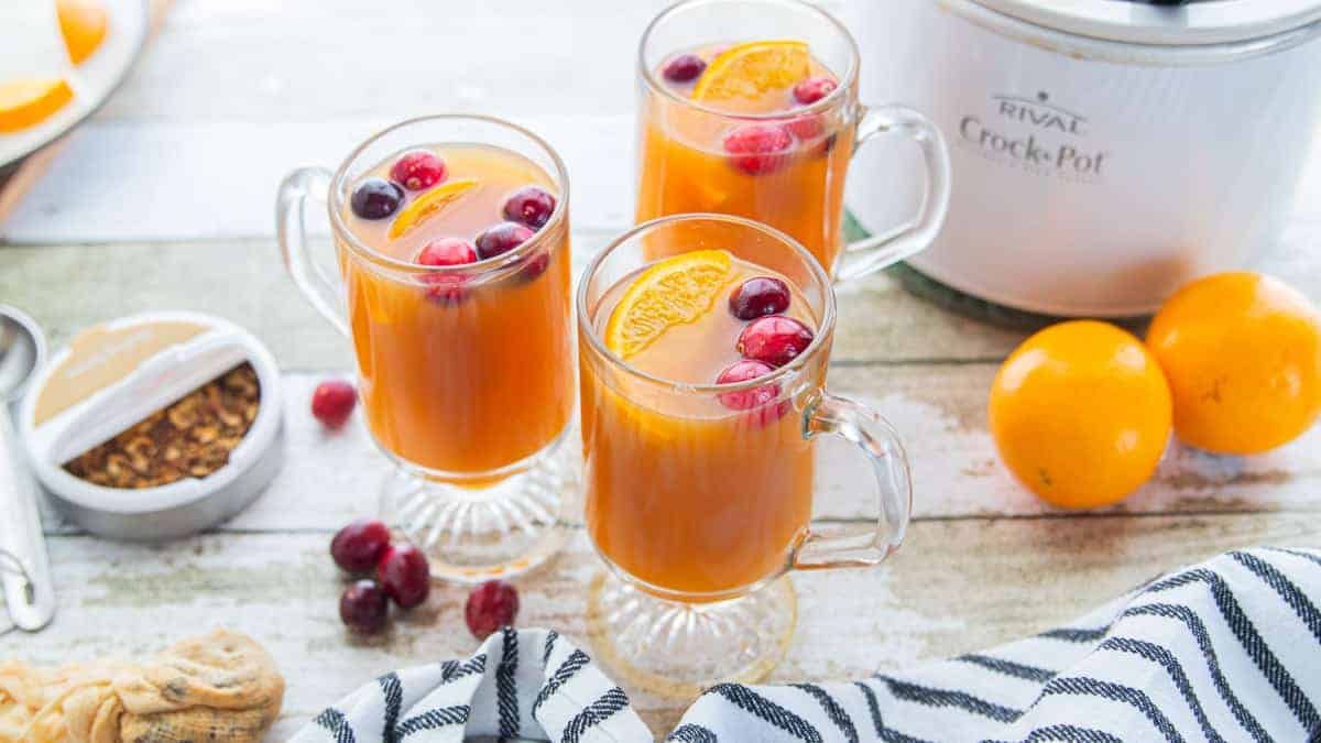  three mugs of apple cider with mulling spices on the side and a crockpot behind the mugs.