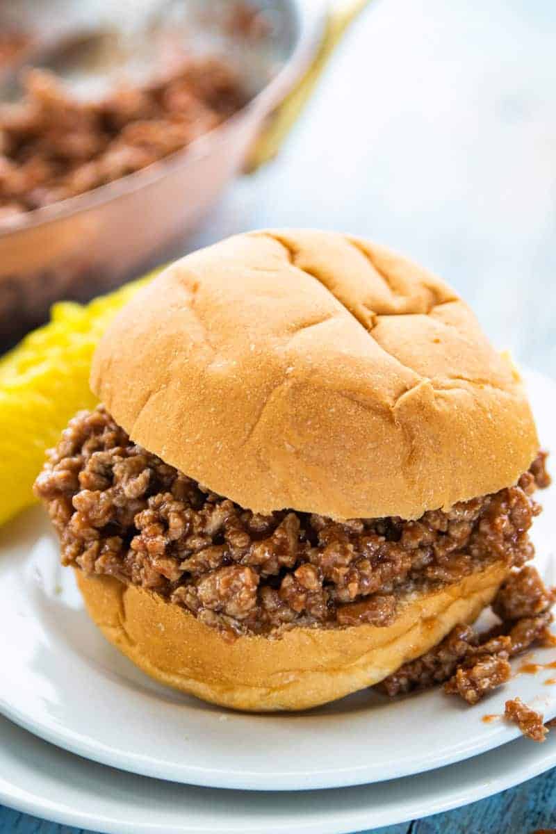 a sloppy Joe sandwich on a white plate with a skillet of sloppy Joe meat behind the plate.