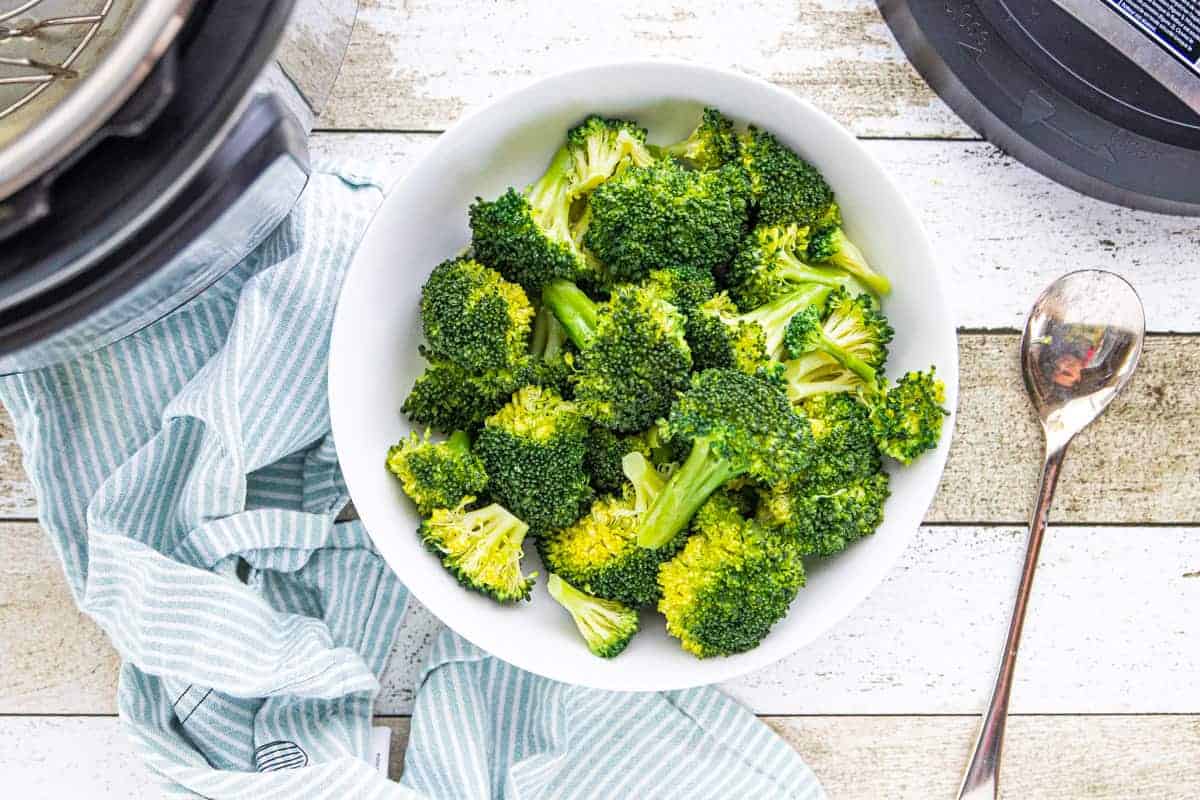 steamed broccoli in a white serving bowl with an instant pot on the side all on top of a wooden surface.